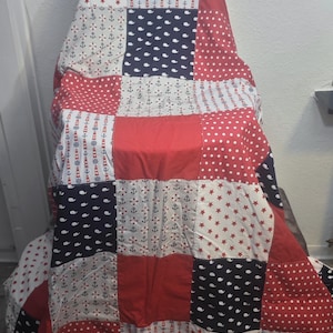 Könnte beinhalten: Ein Patchwork-Quilt mit nautischem Thema, mit roten, weißen und marineblauen Quadraten. Der Quilt zeigt Muster von Ankern, Sternen, Walen und Punkten. Der Quilt ist über einen Stuhl drapiert.