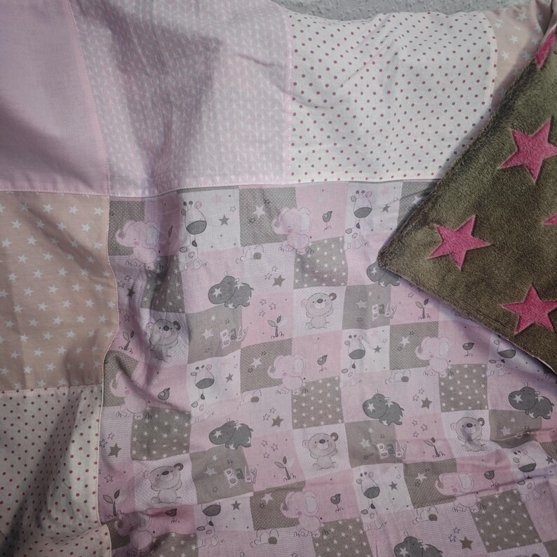 May include: A patchwork baby blanket with pink, tan, and gray squares. The blanket features star and polka dot patterns, along with elephant and bear illustrations. A brown pillow with pink star appliqu&eacute;s is in the corner.