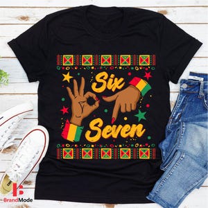 May include: Black t-shirt with a colorful graphic design. The design features the words "Six Seven" in yellow, with hand gestures and decorative elements in red, green, and yellow. The shirt is paired with blue jeans and white sneakers.