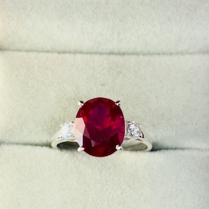 Könnte beinhalten: Ein silberner Ring mit einem großen ovalen Rubin-Edelstein und zwei kleineren runden weißen Edelsteinen auf der Ringschiene.