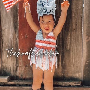 May include: A baby wearing a red, white, and blue crochet outfit with fringe, a blue bow headband, and brown cowboy boots. The baby is holding a small American flag and smiling in front of a wooden fence.