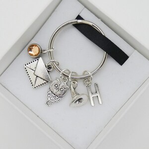 May include: Silver keyring with a silver owl charm, a silver envelope charm, a silver hat charm, and a silver letter H charm. The keyring also has a small orange crystal charm.