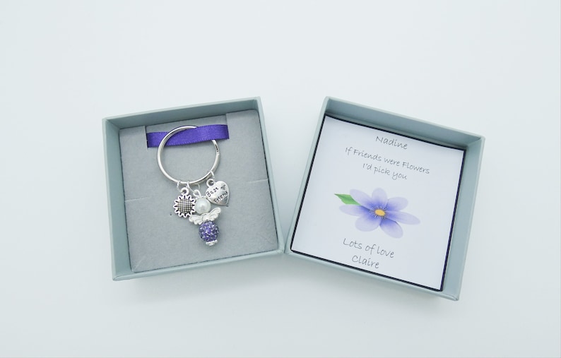 May include: A silver keyring with a purple angel charm, a sunflower charm, and a heart charm that says "Best of Friends". The keyring is in a small gray box with a purple ribbon. The box also contains a card with the text "Nadine, If friends were flowers I'd pick you. Lots of love, Claire."