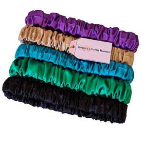 May include: Five satin hair scrunchies in different colors: purple, beige, teal, green, and black. The scrunchies are stacked on top of each other. A pink label with the text "Bonita's Satin Bonnet" is attached to the scrunchies.