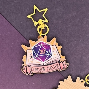 Op de afbeelding: Een houten sleutelhanger met een paarse en roze 20-zijdige dobbelsteen, een boek en de tekst "Dungeon Master". De sleutelhanger heeft een gouden sterbedel.