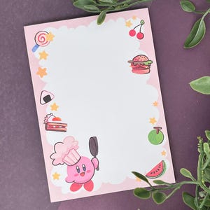 May include: Pink notepad with a white cloud-shaped border. The border is decorated with various cartoon images, including a pink character with a chef's hat, a hamburger, a watermelon, a cherry, a lollipop, a piece of cake, a rice ball, and a green fruit.