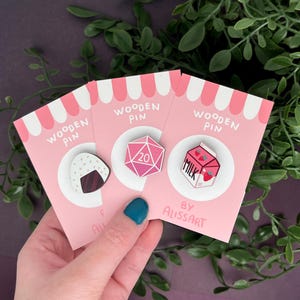 Könnte beinhalten: Drei rosa und weiße Karten mit einem gestreiften Rand. Jede Karte zeigt eine andere Holz-Anstecknadel: ein rosa und weißes Onigiri, ein rosa 20-seitiger Würfel und ein rosa Karton Milch mit Erdbeeren. Die Karten sagen alle "Wooden Pin" und die untere Karte sagt "By Alissart".