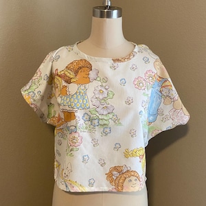 May include: A white, short-sleeved crop top featuring a repeating pattern of cartoon characters, flowers, and bunnies. The top is displayed on a mannequin. The design includes pastel colors such as blue, pink, and yellow.