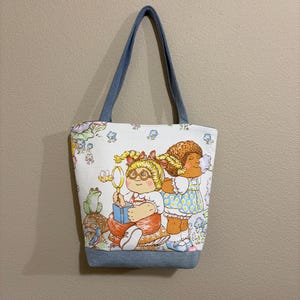 May include: A white tote bag featuring a colorful illustration of two children. The bag has denim blue handles and a denim blue base. The illustration includes a girl with glasses holding a magnifying glass and a girl in a blue dress.