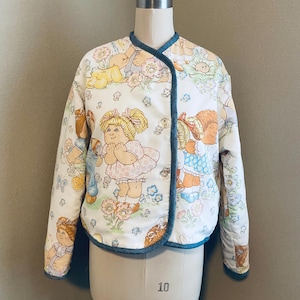 May include: A vintage, quilted jacket featuring a repeating pattern of Cabbage Patch Kids on a cream background. The jacket has a round collar, long sleeves, and a button closure. The trim is a medium blue color.
