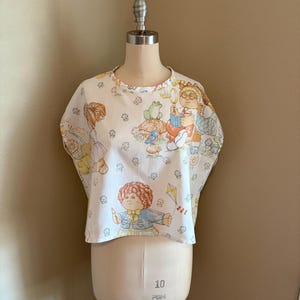 May include: A white short-sleeved top featuring a repeating print of Cabbage Patch Kids characters, including children with yarn-like hair, a frog, a blue bird, and a kite. The fabric has a light-colored background with small floral accents throughout.