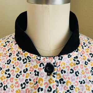 May include: A close-up of a jacket on a mannequin. The jacket features a white background with a colorful leopard print in black, pink, and yellow. It has a black collar and a single black button closure.