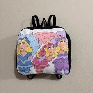 May include: Black and white backpack featuring a colorful graphic of three Muppet characters. The backpack has black straps and a black top. The graphic shows the characters in various outfits, with a light blue, pink, and purple color scheme.
