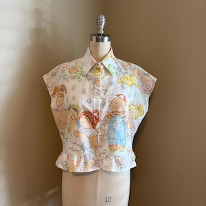 May include: A sleeveless button-down blouse in white fabric featuring a colorful repeating print of Cabbage Patch Kids dolls. The shirt has a pointed collar, a front button placket, and a ruffled hemline at the waist, showcasing various doll characters.
