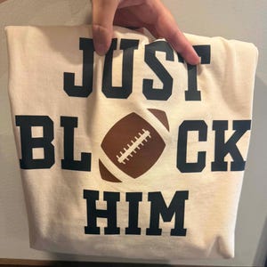 May include: A cream-colored t-shirt featuring the black text "JUST BLOCK HIM" in a bold, athletic font. The word "BLOCK" is split by a brown and white graphic illustration of an American football positioned in the center of the shirt.