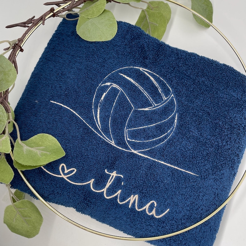 May include: A navy blue towel with a white embroidered volleyball design and the name "ertina" in cursive. The towel is folded and displayed with a gold hoop and artificial greenery.