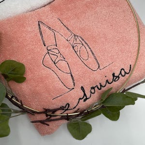 May include: A pink towel embroidered with a black outline of ballet shoes and the name "Louisa" in cursive. The towel is partially framed by a gold-colored metal ring and decorated with green leaves.