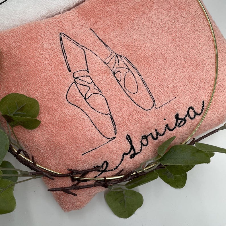 May include: A pink towel embroidered with a black outline of ballet shoes and the name "Louisa" in cursive. The towel is partially framed by a gold-colored metal ring and decorated with green leaves.