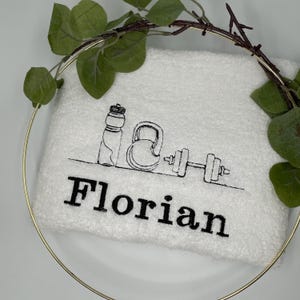 May include: White fitness towel embroidered with black line art of a water bottle, kettlebell, and dumbbell. The name "Florian" is embroidered in bold black letters below the illustrations. The towel is framed by a gold ring and greenery.