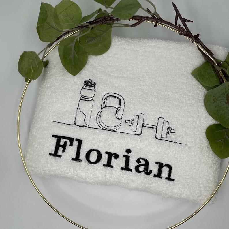May include: White fitness towel embroidered with black line art of a water bottle, kettlebell, and dumbbell. The name "Florian" is embroidered in bold black letters below the illustrations. The towel is framed by a gold ring and greenery.