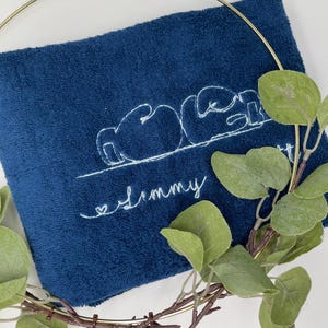 May include: A navy blue towel with white embroidered text and a boxing glove design. The word "Jimmy" is embroidered in cursive below the design. A gold-colored ring and green leaves partially frame the towel.