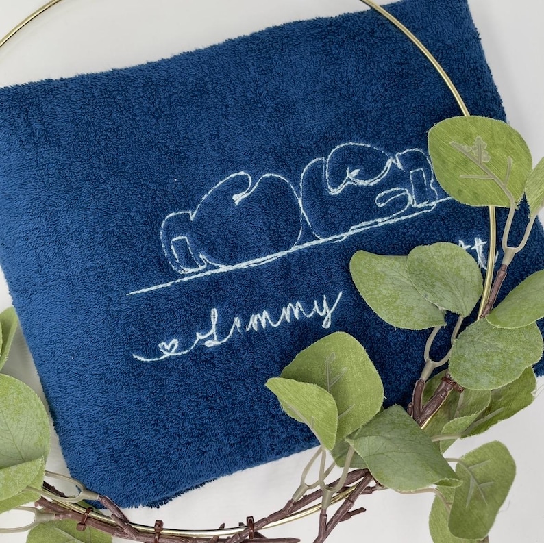 May include: A navy blue towel with white embroidered text and a boxing glove design. The word "Jimmy" is embroidered in cursive below the design. A gold-colored ring and green leaves partially frame the towel.