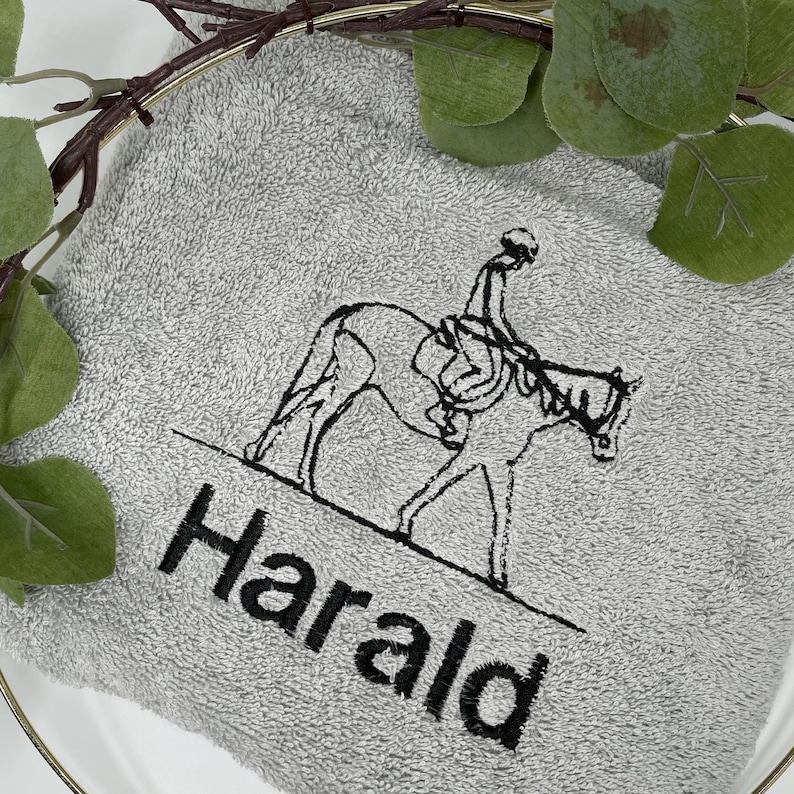May include: A gray towel with the name "Harald" embroidered in black. The towel features a black line drawing of a person riding a horse. The towel is surrounded by greenery and a gold-colored ring.