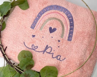 May include: A pink plush towel with an embroidered rainbow and the name "Pia" in purple thread. The rainbow has pink, green, and purple stripes.