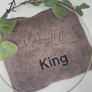 May include: A brown towel with a black embroidered barbell design and the word "King". The towel is surrounded by a gold ring and green leaves.