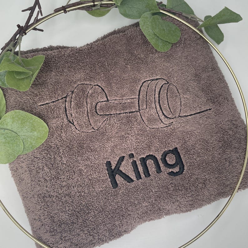 May include: A brown towel with a black embroidered barbell design and the word "King". The towel is surrounded by a gold ring and green leaves.