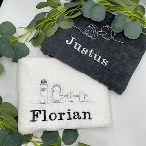 May include: Two folded gym towels, one charcoal gray and one white, with embroidered names and fitness-themed line art. The gray towel has the name "Justus" and a dumbbell design. The white towel has the name "Florian" and a water bottle, kettlebell, and dumbbell design.