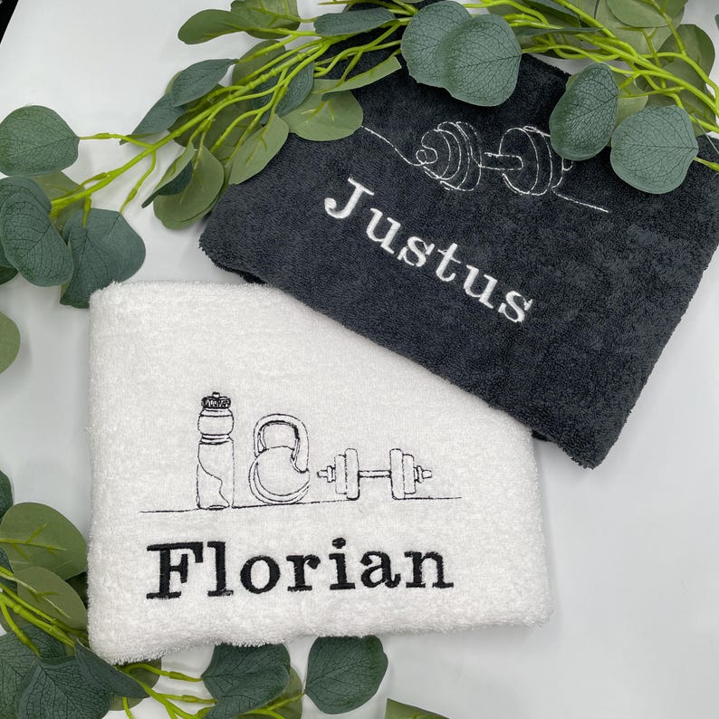 May include: Two folded gym towels, one charcoal gray and one white, with embroidered names and fitness-themed line art. The gray towel has the name "Justus" and a dumbbell design. The white towel has the name "Florian" and a water bottle, kettlebell, and dumbbell design.