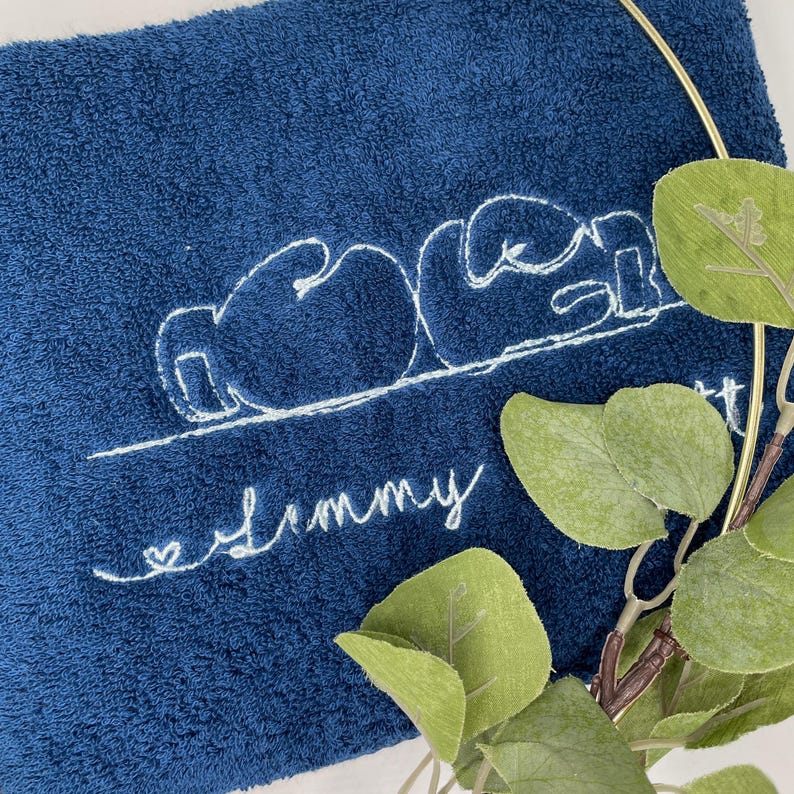 May include: A navy blue towel with white embroidered text and a boxing glove design. The word "Mom" is above the name "Jimmy" in cursive, with a small heart. Green leaves and a gold ring are in the background.