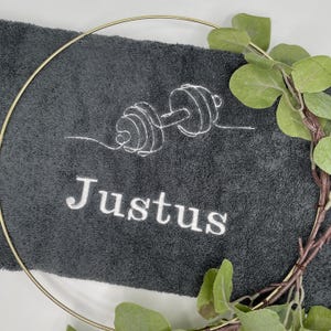 May include: A dark gray gym towel embroidered with a white outline of a barbell and the name "Justus". A gold-colored circular frame and a sprig of green leaves partially surround the towel.