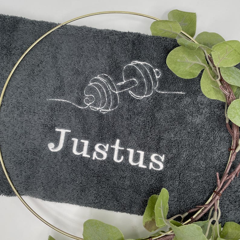 May include: A dark gray gym towel embroidered with a white outline of a barbell and the name "Justus". A gold-colored circular frame and a sprig of green leaves partially surround the towel.