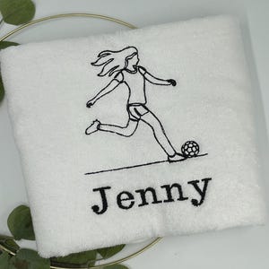 May include: White towel with a black embroidered soccer player kicking a soccer ball. The name "Jenny" is embroidered in black below the soccer player.