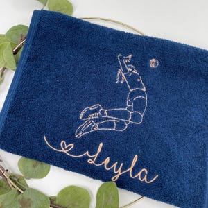 May include: Navy blue towel with an embroidered volleyball player in action and a volleyball. The name "Leyla" is embroidered in a cursive font. The towel is folded and placed on a gold ring with green leaves.