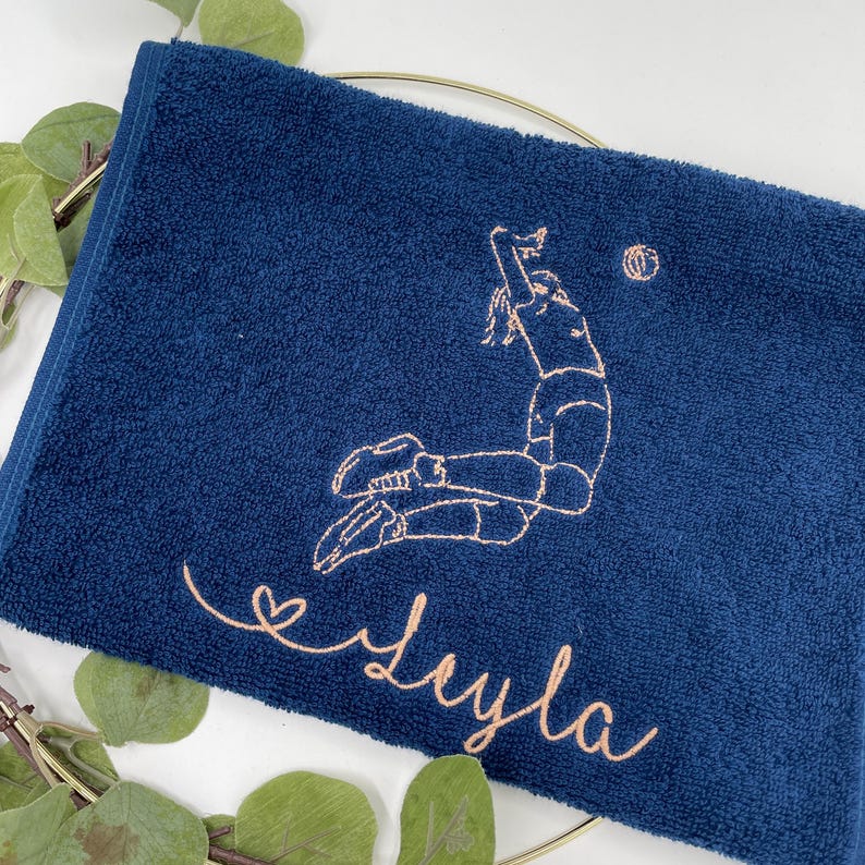 May include: Navy blue towel with an embroidered volleyball player in action and a volleyball. The name "Leyla" is embroidered in a cursive font. The towel is folded and placed on a gold ring with green leaves.