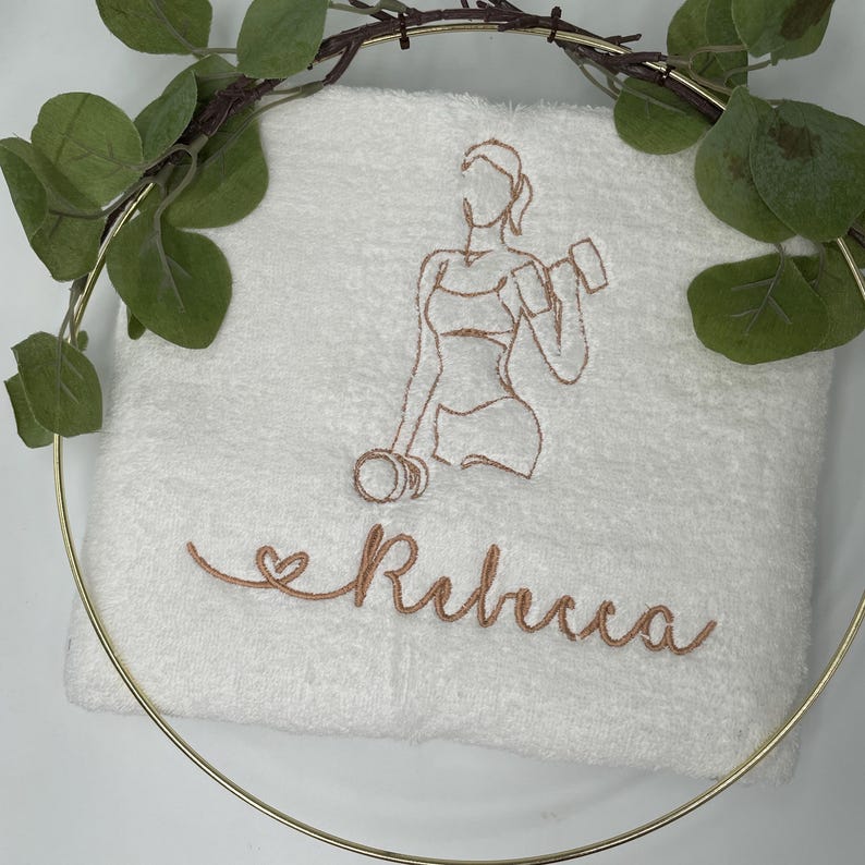 May include: White fitness towel embroidered with a copper-colored outline of a woman lifting weights. The name "Rebecca" is embroidered in a cursive font below the image. The towel is decorated with greenery and a gold hoop.