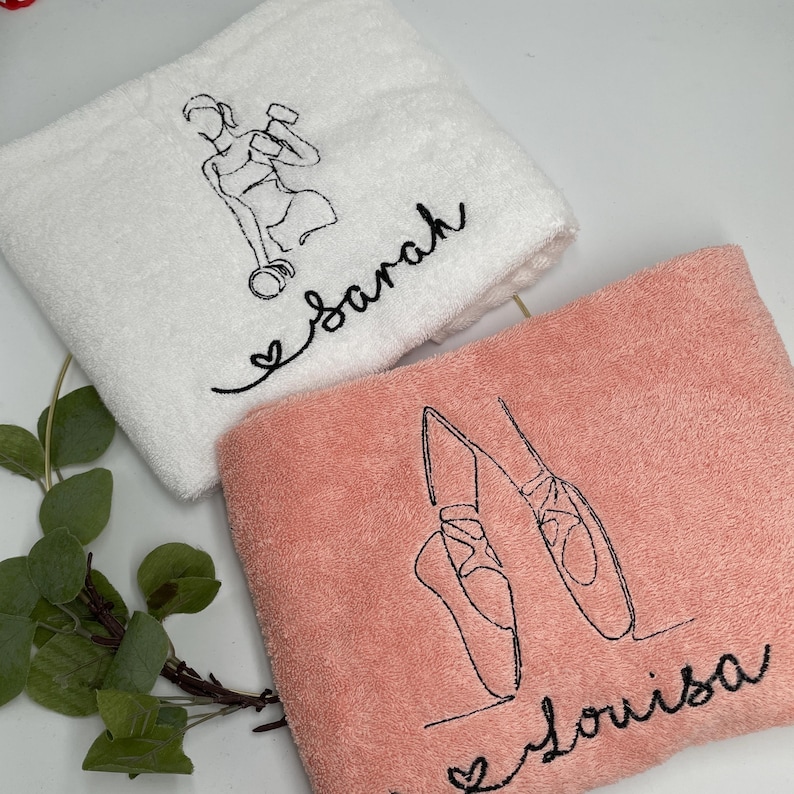 May include: Two embroidered towels. One white towel features a black line drawing of a person lifting weights, with the name "Sarah" in cursive. The other is a pink towel with a ballet shoe design and the name "Louisa".