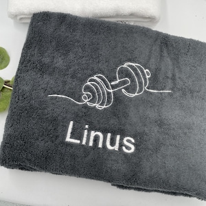 May include: A dark gray towel with a white embroidered dumbbell design and the name "Linus". A white towel is visible in the background. The towel is likely made of a soft, absorbent material, suitable for use after a workout.