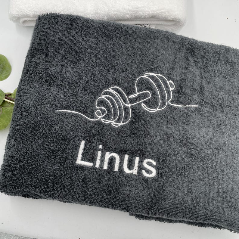 May include: A dark gray towel with a white embroidered dumbbell design and the name "Linus". A white towel is visible in the background. The towel is likely made of a soft, absorbent material, suitable for use after a workout.