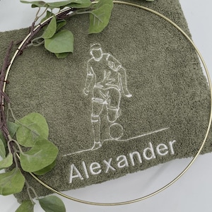 May include: A green towel with a white embroidered soccer player and the name "Alexander". A gold metal ring and faux greenery frame the towel. The towel is a soft, textured material.