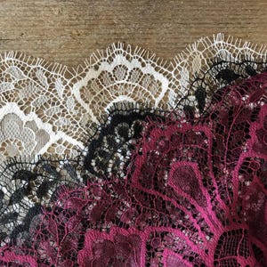 Può includere: Primo piano di tre diversi tessuti di pizzo. Il pizzo è bianco, nero e magenta intenso. Il pizzo presenta un delicato motivo floreale con bordi smerlati. Lo sfondo è una superficie di legno marrone chiaro.