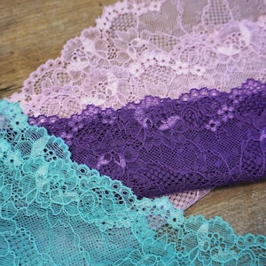 May include: Close-up of three pieces of lace fabric in shades of teal, purple, and pink. The lace has a delicate floral pattern.