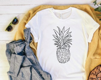 Pode incluir: Uma camiseta branca com um desenho de linha preta de um abacaxi na frente. A camiseta está sobre uma superfície de madeira branca com um par de calças jeans azuis, um casaco amarelo e um par de sapatos marrons de pele de cobra.