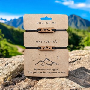 Puede incluir: Un par de pulseras de cuerda negra con colgantes de madera en forma de montaña. Las pulseras están empaquetadas en una tarjeta con el texto "ONE FOR ME" y "ONE FOR YOU" y el mensaje "My heart and I agree that you are the only one for me."