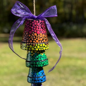 May include: A stack of five painted terracotta pots, suspended by a twine string and adorned with a purple ribbon. Each pot is painted with a different color, transitioning from red to purple, and covered in a dot pattern.