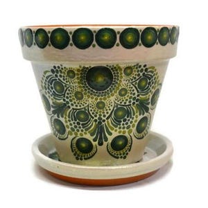 May include: A hand-painted terracotta flower pot with a matching saucer. The pot features a cream-colored base with a detailed green mandala design and a border of green circles. The rim and base are terracotta.