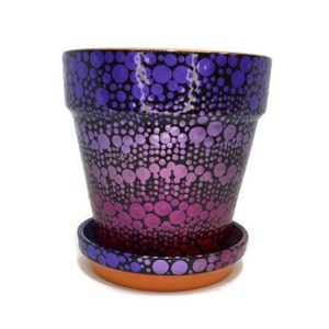 May include: A terracotta flower pot with a matching saucer, hand-painted with a purple and pink ombre dot pattern.